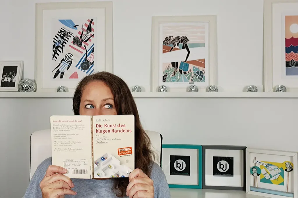 Britta hält sich das Buch "Die Kunst des klugen Handelns" vor das Gesicht und schaut darüber verschmitzt nach rechts, während sie provokant fragt: Die Wahrheit über den DIY Website Trend – verschwendest Du als Selbstständige Deine wertvollste Ressource?
