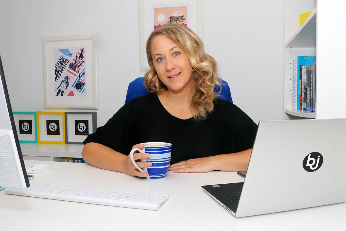 Britta sitzt an ihrem Schreibtisch hinter ihrem Laptop, hält eine Tasse Galão in der Hand und lächelt in die Kamera als Symbolbild für "So kannst Du mit mir zusammenarbeiten".