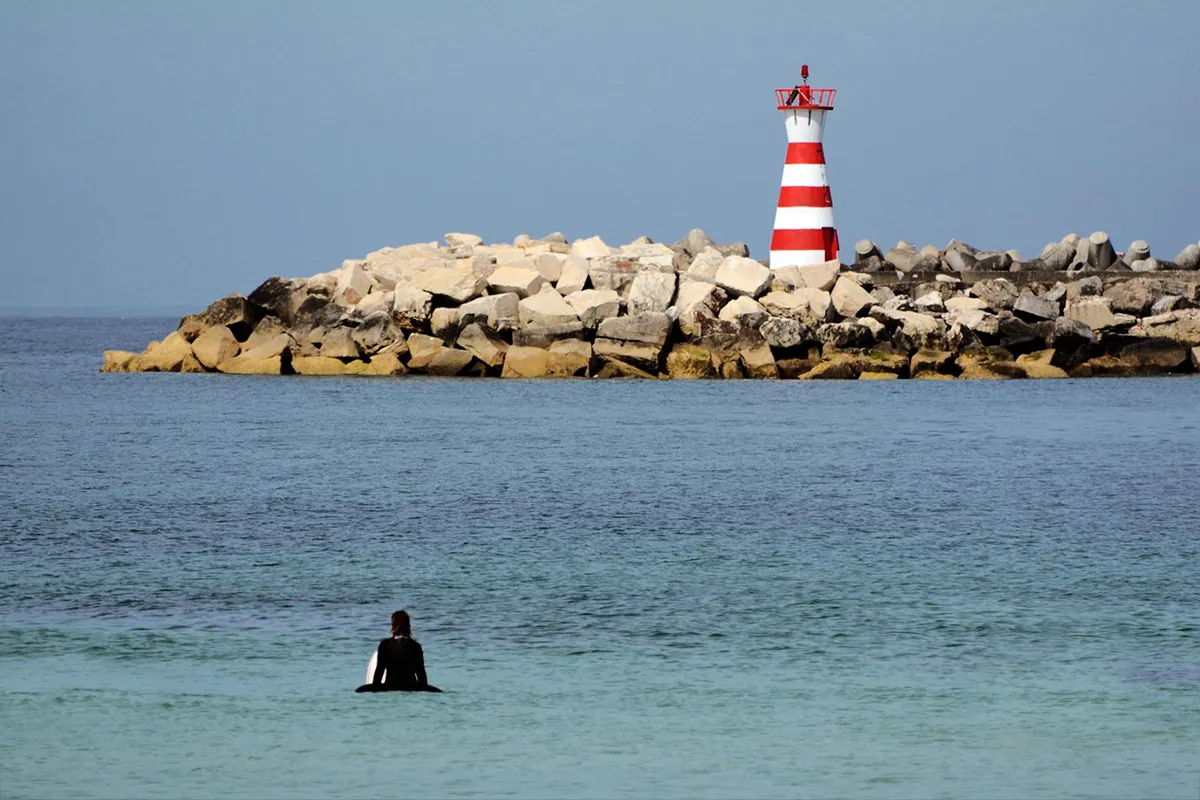 Britta sitzt anlässlich ihres Portuversary auf ihrem Surfboard vor einem Leuchtturm und wartet auf die nächste Welle.