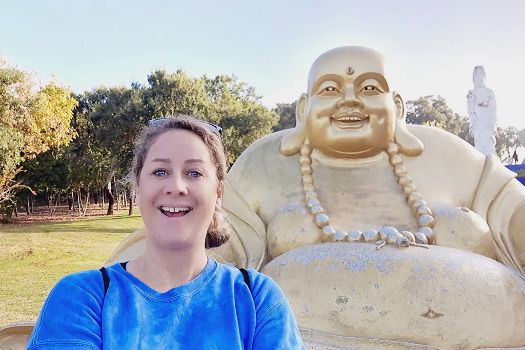 Britta lacht mit einer dickbäuchigen, goldenen Buddha Figur um die Wette im Buddha Eden Garten in Portugal.